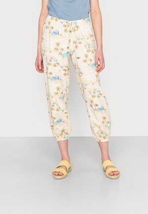 Personne portant un pantalon de jogging crème avec un imprimé plage et palmier, un haut bleu clair et des sandales jaunes, debout à l'intérieur.