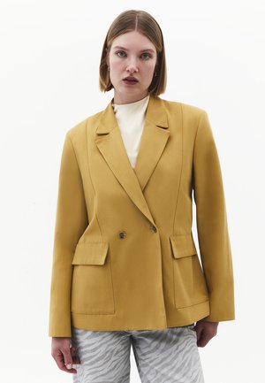 Kvinde med kort hår iført en sennepsgul dobbeltradet blazer og lysegrå zebramønstrede bukser mod en ensfarvet hvid baggrund.