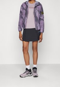 Fialově vzorovaná lehká bunda přes světle fialovou košili a tmavou sukni. Doplňena šedými a růžovými turistickými botami a bílými ponožkami.