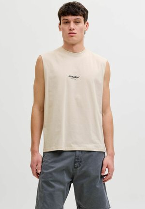 Noor mees kannab beežil värvilist varrukateta T-särki kirjaga "JJ Studios" ning tumedat halli lühikest pükst, seistes ühetaustal heleda tausta ees.