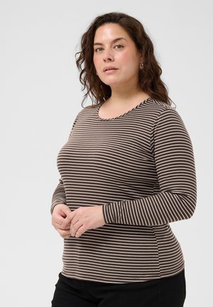 Femme aux cheveux bruns bouclés portant une chemise à rayures marron et blanc à manches longues et un pantalon noir, debout devant un fond uni.