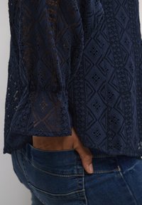 Blusa lunga blu navy con texture in pizzo geometrico intricata e arricciature ai polsini, abbinata a jeans in denim blu.