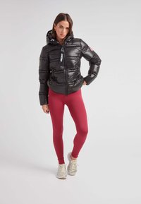 Svart vadderad jacka med huva, dragkedja och logodetalj, kombinerad med röda ribbade leggings och ljusa sneakers. Slät textur, åtsittande design.