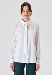 Blusa bianca con maniche lunghe, colletto e dettaglio a cravatta. Presenta un tessuto liscio e trasparente con una chiusura frontale con bottoni e polsini a sbuffo.