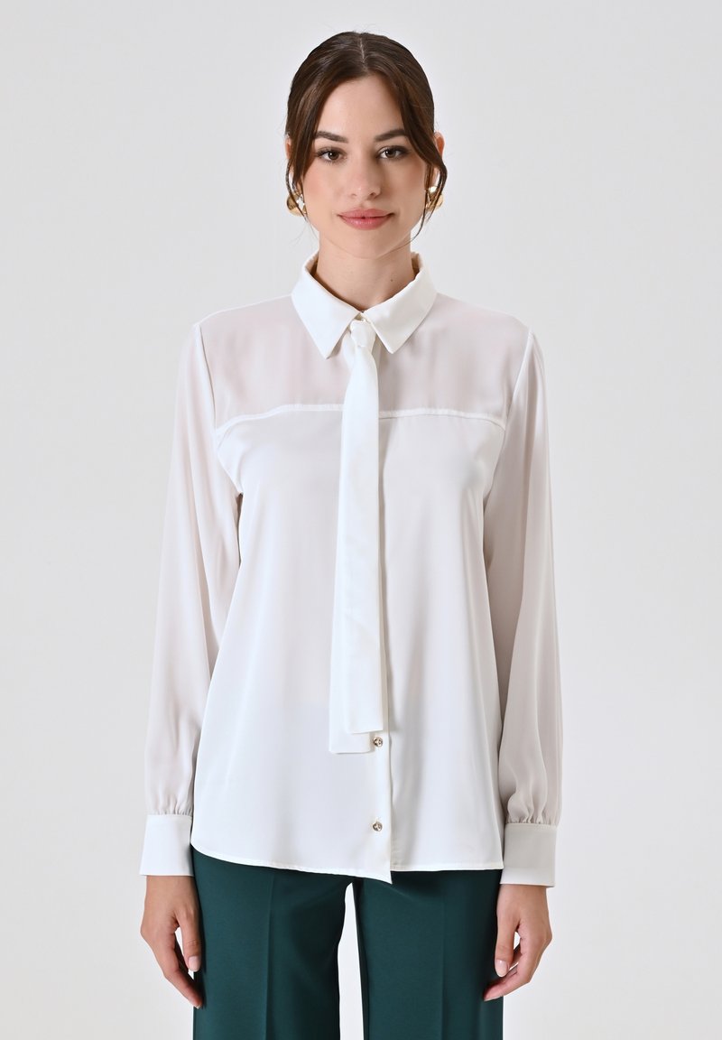 Blusa bianca con maniche lunghe, colletto e dettaglio a cravatta. Presenta un tessuto liscio e trasparente con una chiusura frontale con bottoni e polsini a sbuffo.