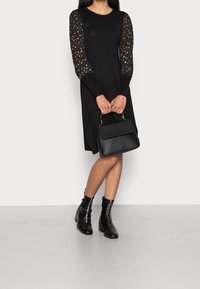 Robe noire avec de longues manches texturées ornées de pois dorés. Le mannequin tient un sac à main en cuir noir, associé à des bottines, sur un fond gris.