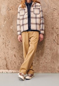 Rutig, långärmad jacka i beige, bruna och blå nyanser, med två bröstfickor. I kombination med ljusbruna, löst sittande byxor och bruna sneakers.