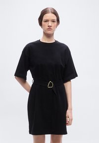 Γυναίκα με καστανά μαλλιά, φορώντας μαύρο φόρεμα με ζώνη και κοντά μανίκια, στέκεται μπροστά σε απλό φωτεινό φόντο, στραμμένη προς τα εμπρός με ουδέτερη έκφραση.