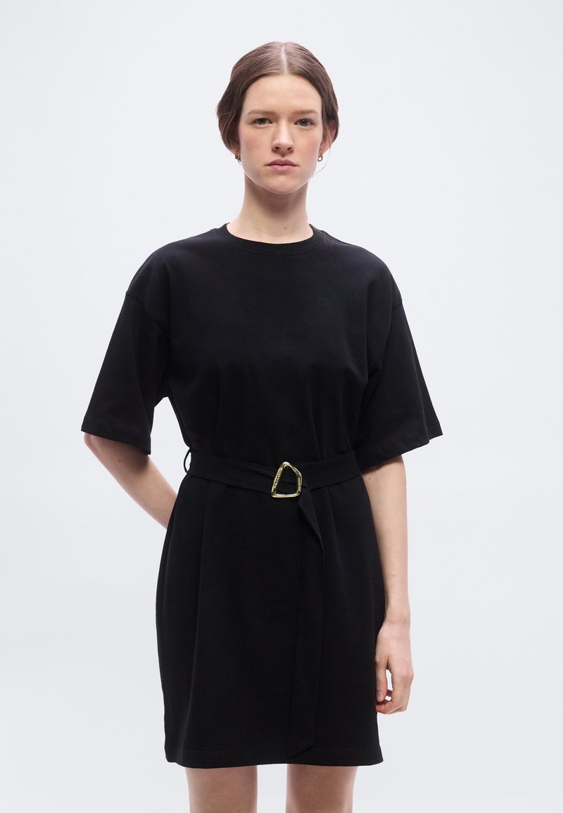 Γυναίκα με καστανά μαλλιά, φορώντας μαύρο φόρεμα με ζώνη και κοντά μανίκια, στέκεται μπροστά σε απλό φωτεινό φόντο, στραμμένη προς τα εμπρός με ουδέτερη έκφραση.