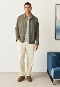 Giacca verde oliva a bottoni con un motivo testurizzato, t-shirt grigia sotto, pantaloni color crema e sneaker in suede marrone.