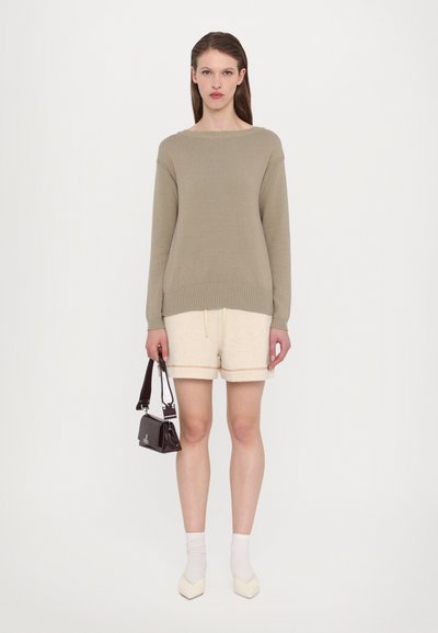 Μπεζ πλεκτό πουλόβερ με φαρδιά λαιμόκοψη, κρεμ σορτσάκια με υφή, λευκές κάλτσες και μια μικρή γυαλιστερή σκούρα τσάντα.