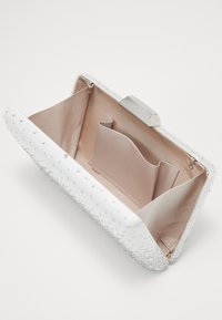 Biała kopertówka wykonana z teksturowanego materiału, z srebrnymi akcentami i ozdobami, z beżową podszewką i wewnętrzną kieszenią.