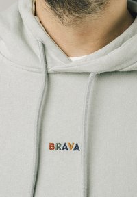 Světle šedá mikina s kapucí a stahovací šňůrkou, na levém prsu má vícebarevnou výšivku "BRAVA". Měkká, texturovaná tkanina.