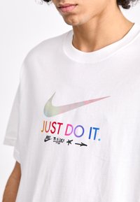 Vit t-shirt i bomull med en färgglad gradient av Nike swoosh och texten "JUST DO IT." Svarta detaljer inkluderar "NIKE B. SIDES" och enkla grafiska element.