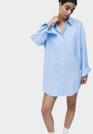 Kvinde iført en oversized lyseblå skjortekjole med opkrogede ærmer, der står foran en ensfarvet hvid baggrund.