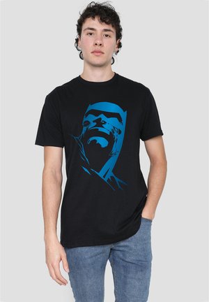 Young man stands with one hand in pocket wearing black t-shirt with blue stylized Batman face and blue jeans against plain background.