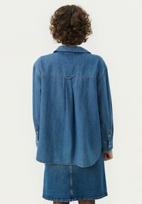 Camicia in denim blu con vestibilità rilassata, colletto a punta, maniche lunghe, polsini con bottone e orlo arrotondato. Il retro presenta una piega centrale.