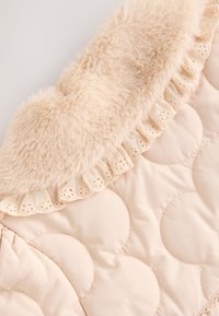 Veste matelassée beige clair avec col en fausse fourrure et dentelle en bordure. Présente des motifs ronds matelassés et une texture douce partout.