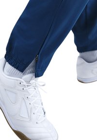 Blauwe sportbroek met een ritsdetail bij de enkel, gecombineerd met witte sneakers met gaasaccenten en een gumrubberen zool.