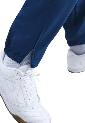 Blaue Sporthosen mit einem Reißverschlussdetail am Knöchel, kombiniert mit weißen Sneakern, die Netzapplikationen und eine Gummisohle haben.
