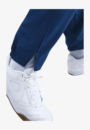 Blauwe sportbroek met een ritsdetail bij de enkel, gecombineerd met witte sneakers met gaasaccenten en een gumrubberen zool.
