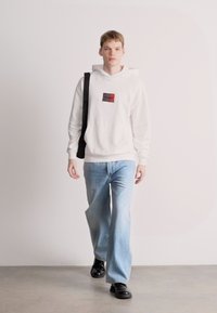 Λευκή κουκούλα με μικρή κόκκινη και μαύρη ετικέτα, ανοιχτό μπλε τζιν, και μαύρα παπούτσια. Το μοντέλο στέκεται μπροστά σε ουδέτερο φόντο.