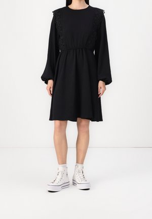 Kobieta w czarnej, długiej sukience do kolan z koronkowymi detalami na ramionach, w połączeniu z białymi, wysokimi trampkami i skarpetkami.