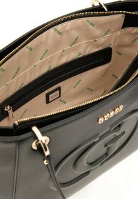 Sac à main en simili cuir noir avec fermeture éclair dorée et logo en relief. L'intérieur est doublé de beige avec un motif "GUESS" répété et des poches.