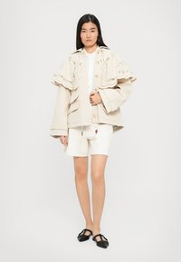 Tânără îmbrăcată într-o jachetă oversized bej cu o capă din dantelă, pantaloni scurți albi și pantofi negri cu vârf ascuțit, stând în fața unui fundal alb.