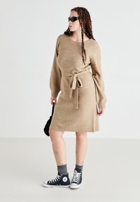 Vestido de punto beige con cinturón de lazo, mangas largas y un corte relajado. Combinado con zapatillas altas negras y calcetas grises.