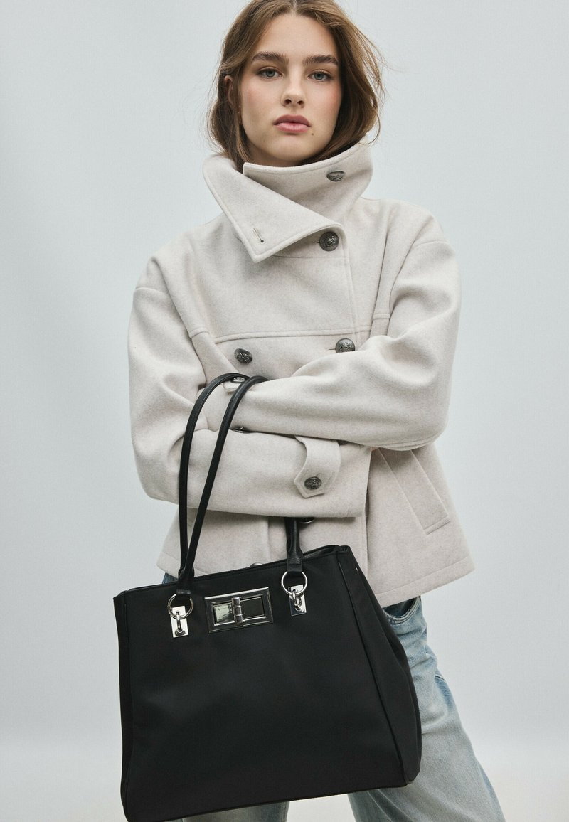 Jeune femme aux cheveux châtain clair portant un manteau beige clair boutonné et tenant un grand sac à main noir sur un fond uni.