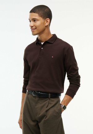 Junger Mann mit einem dunkelbraunen langärmligen Poloshirt, karierten braunen Hosen mit einem schwarzen Gürtel und einer Armbanduhr, der mit einer Hand in der Tasche steht.