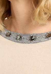 Beżowy sweter z szarym dekoltem ozdobionym diamentowatymi czarnymi kamieniami i srebrnymi akcentami, prezentujący miękką, gładką teksturę.