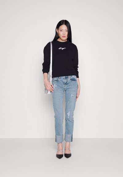 Sweatshirt noir avec logo blanc, jean bleu déchiré et talons aiguilles noirs. Le modèle porte un sac bandoulière argenté, se tenant devant un fond clair.