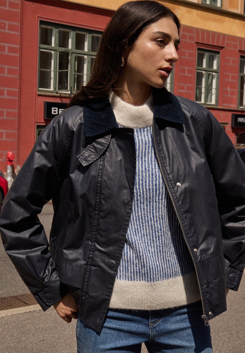 Marinfärgad bombarjacka med en manchesterkrage; kombinerad med en krämfärgad och blå randig tröja; bakgrunden visar färgglada byggnader och en gata.