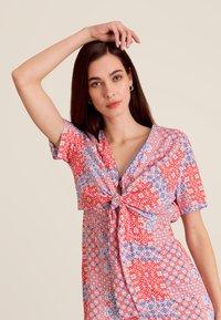 Robe à manches courtes avec des motifs rouges, bleus et blancs, dotée d'un détail noué à l'avant. Tissu léger avec un motif floral et géométrique.