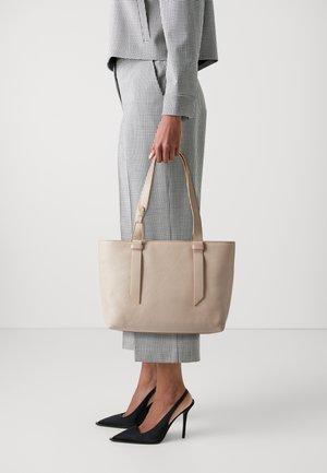 Borsa tote in pelle beige con doppi manici, caratterizzata da una superficie testurizzata e un design minimalista. Indossata con un completo a scacchi e tacchi neri.
