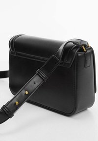 Musta nahkainen olkalaukku, jossa on jämäkkä muotoilu, litteä yläosa, säädettävä hihna kulta- ja metalliosilla sekä minimalistiset yksityiskohdat.