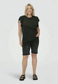 Γυναίκα που χαμογελά, φορώντας μαύρη μπλούζα με φουστίτσα, μαύρες μινι σορτς και μαύρα σανδάλια, στέκεται ενάντια σε έναν απλό φωτεινό φόντο.