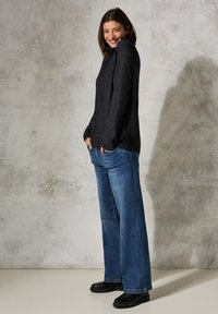 Maglione nero a maglia con maniche lunghe, abbinato a jeans a gamba larga blu chiaro e stivaletti neri, su uno sfondo grigio chiaro con texture.