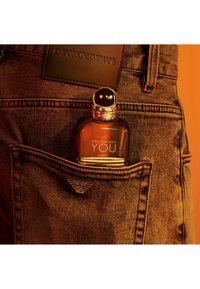 Flacon de parfum Amber étiqueté "Stronger With You" glissé dans la poche arrière d'un jean marron Emporio Armani avec détails du logo.