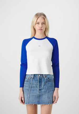 Valkoinen crops-top, jossa on pitkät siniset hihat, pyöreä pääntie ja pieni sininen-logo rinnan kohdalla. Yhdistetty farkkuminihameeseen.