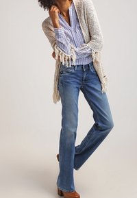 Lichtblauwe flared jeans van denim, gecombineerd met een crèmekleurige gebreide cardigan met franjes. Daaronder een wit-blauw gestreepte blouse.