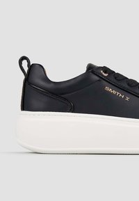 Sneaker in pelle nera con una spessa suola bianca, caratterizzato da dettagli dorati e un design con lacci. Include un tassello sul tallone per indossarle facilmente.