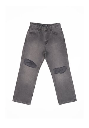 Młody mężczyzna stoi twarzą do przodu, ubrany w czarną bluzę z kapturem i zamkiem, szary t-shirt z nadrukiem, podarte szare dżinsy oraz czarno-białe trampki na jednolitym tle.