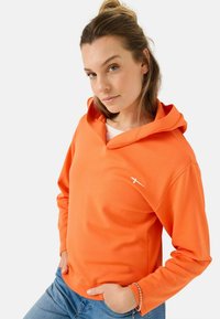 Oranje hoodie van zacht materiaal, met een V-hals ontwerp en een witte logo accent op de borst. G gedragen met blauwe jeans.
