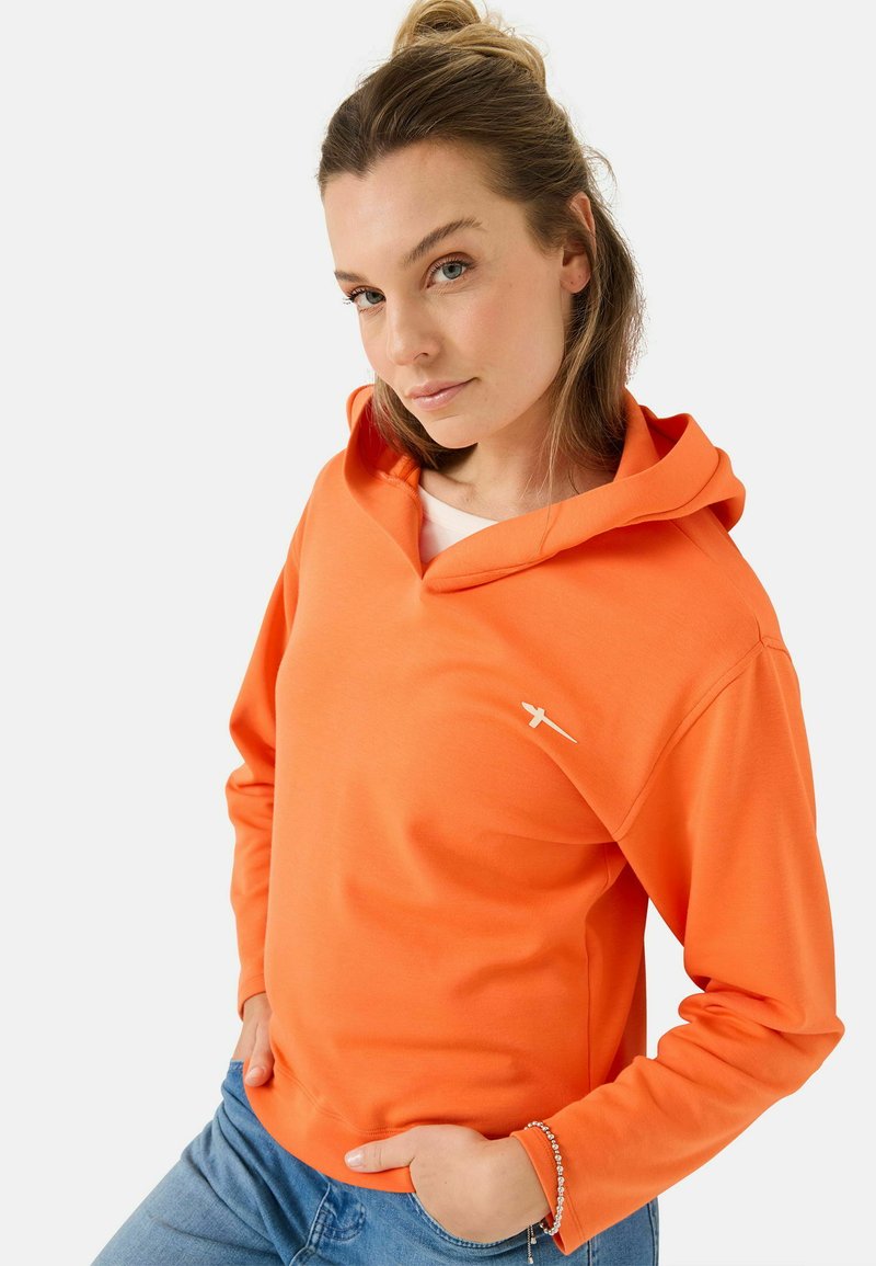 Oranje hoodie van zacht materiaal, met een V-hals ontwerp en een witte logo accent op de borst. G gedragen met blauwe jeans.