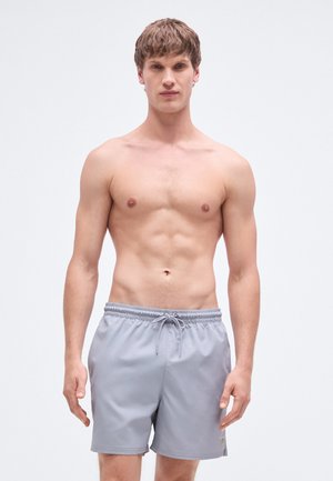 Shorts de bain gris avec une ceinture élastique et un cordon de serrage, présentant une texture lisse et un subtil accent de logo sur l'ourlet.