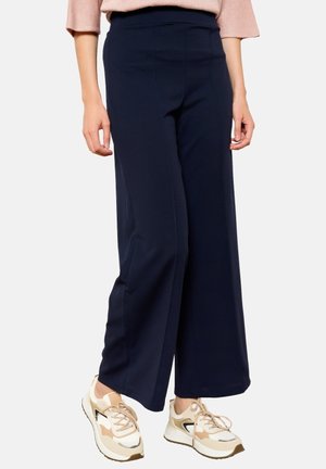 Femme portant un pantalon large bleu marine avec des baskets beiges et blanches, debout devant un fond clair uni.