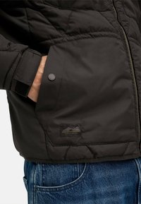 Sort isoleret jakke med quiltet design, der har en trykknap-lukning lomme og et logo-patch på kanten, parret med blå denim jeans.